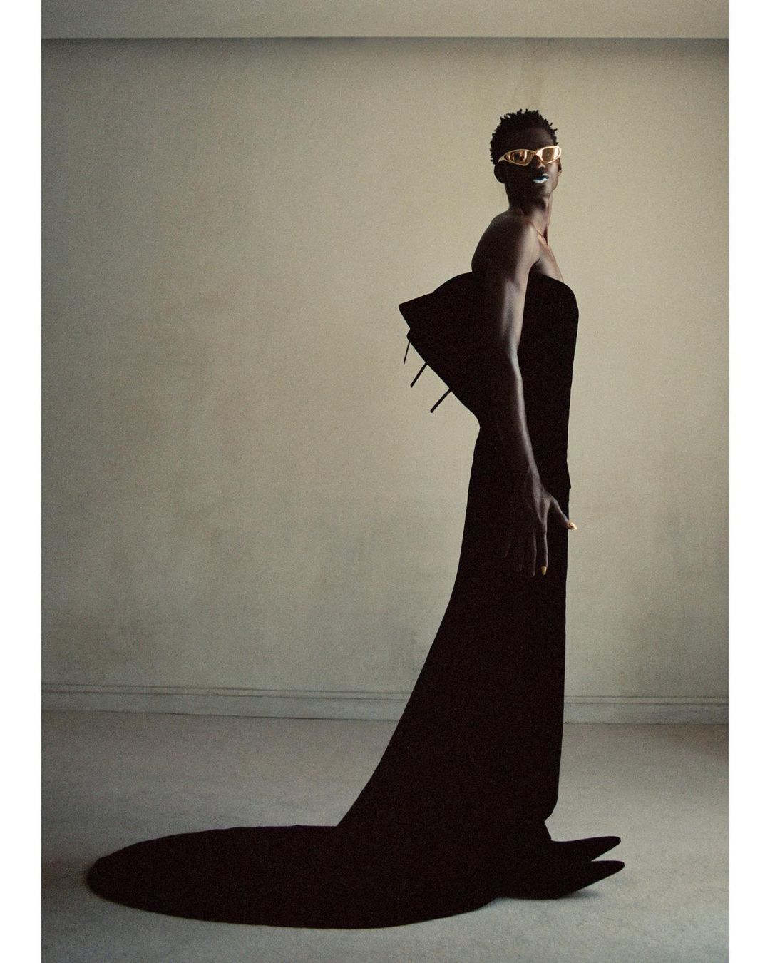 Androgynous fashion model in a long black dress and gold sunglasses poses in a minimalist studio with neutral wall color.