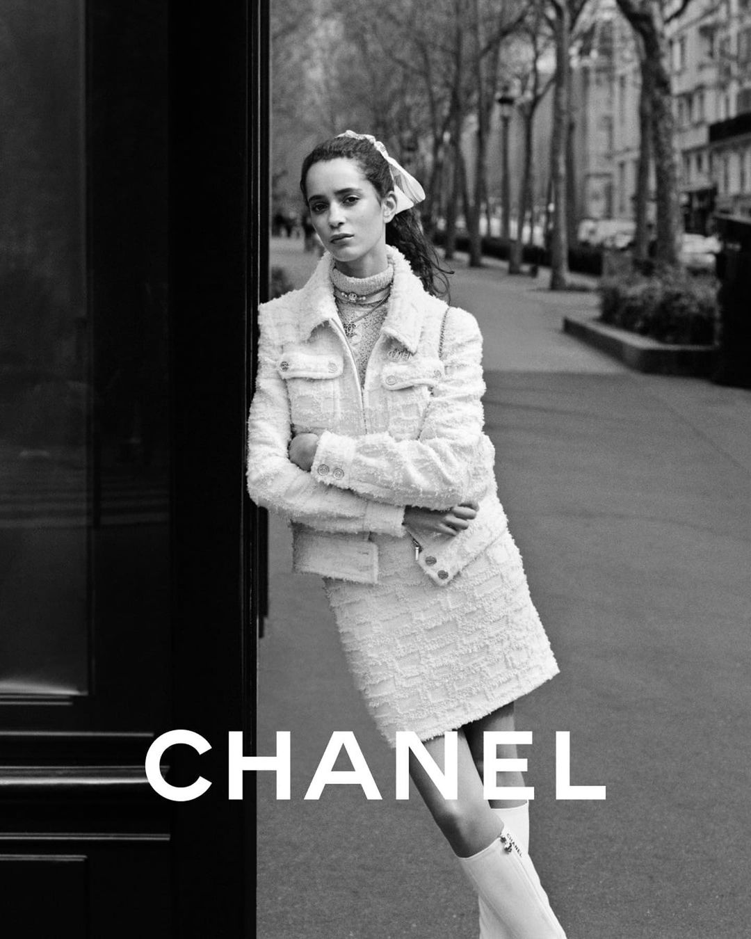 Black and white fashion shot of a young adult woman in a Chanel tweed skirt suit standing in an urban environment near a black door.