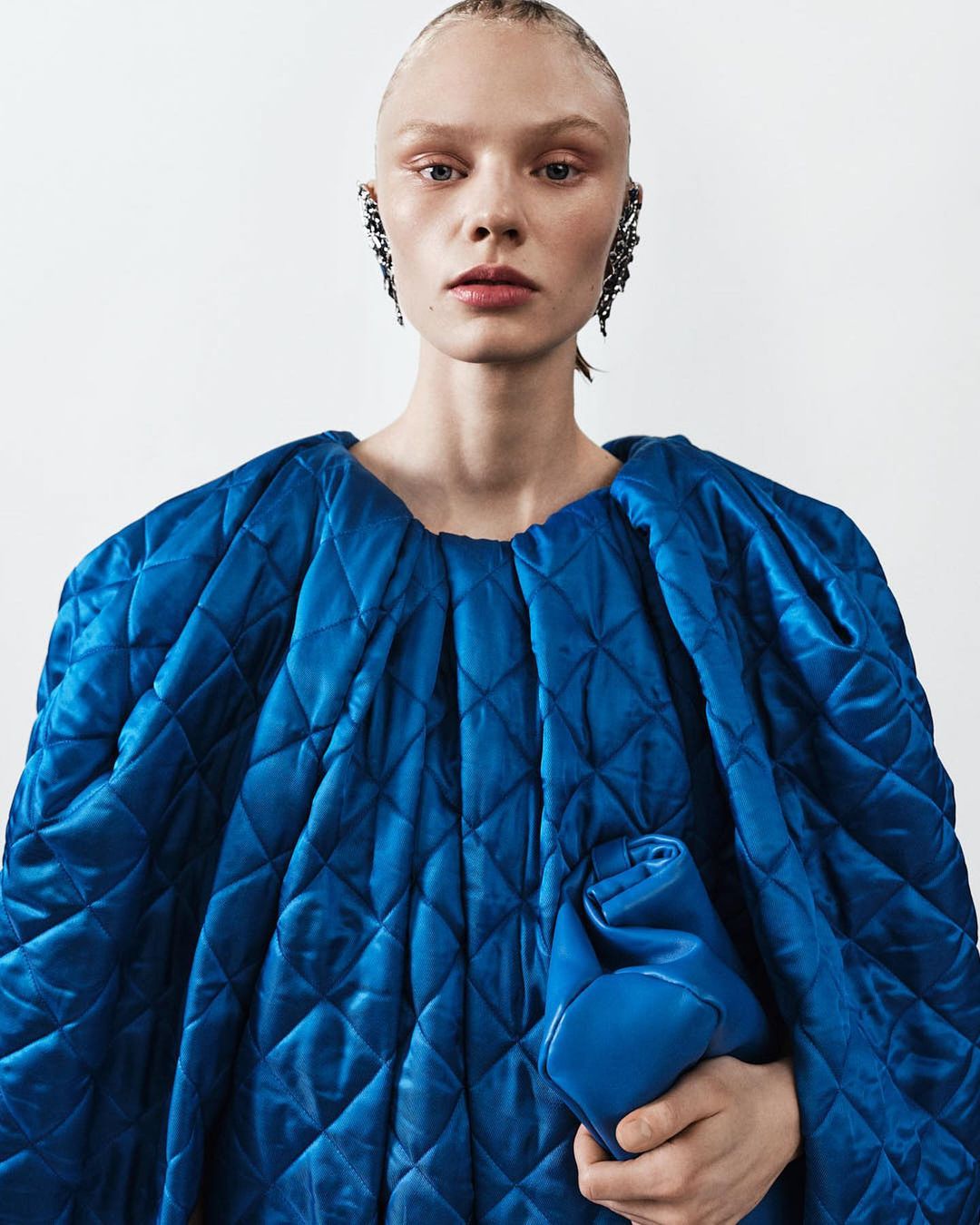 Portrait of a fair-skinned young woman in a blue quilted jacket, holding a matching blue clutch, against a neutral background.