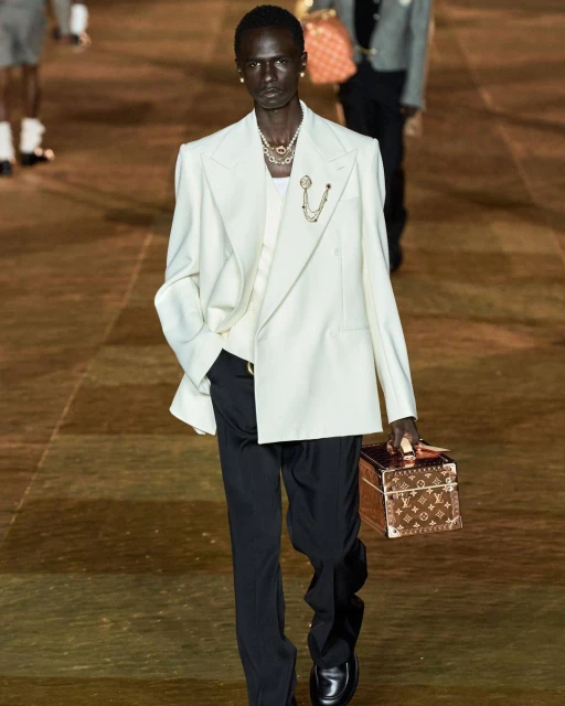 A runway model with dark skin walks in a cream-colored jacket, black pants and boots, and carries a brown-and-gold purse. 