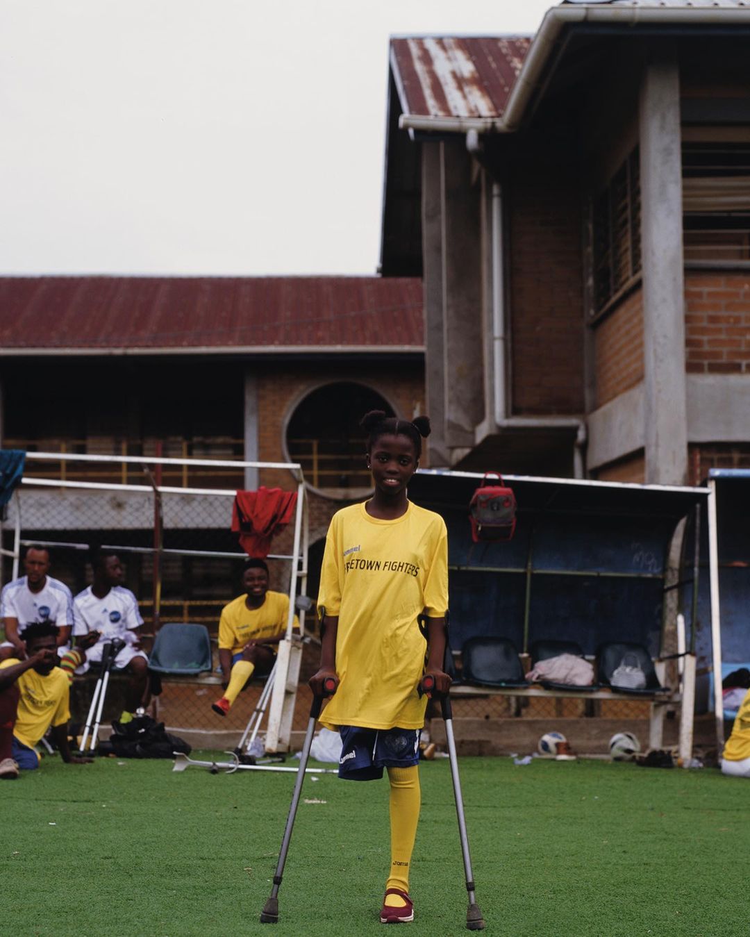 Young disabled female soccer player standing on crutches on a green sports field with a team, building, and overcast sky.