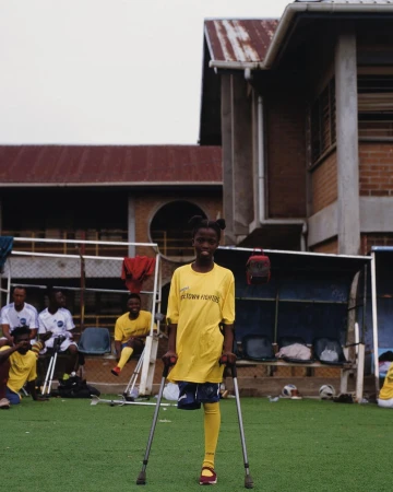 Young disabled female soccer player standing on crutches on a green sports field with a team, building, and overcast sky.