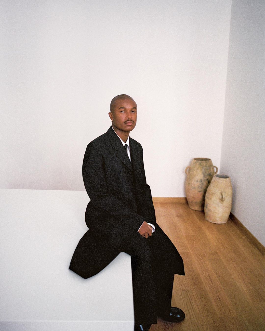 An adult man in a black suit sits on a white platform in a minimally decorated room with a hardwood floor and neutral-toned decorative vases.