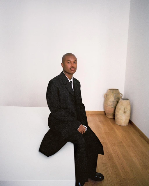 An adult man in a black suit sits on a white platform in a minimally decorated room with a hardwood floor and neutral-toned decorative vases.