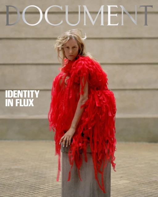 Blonde woman wearing a red, feathery dress poses on a concrete cylinder against a gray wall for a magazine cover titled 'Document'.