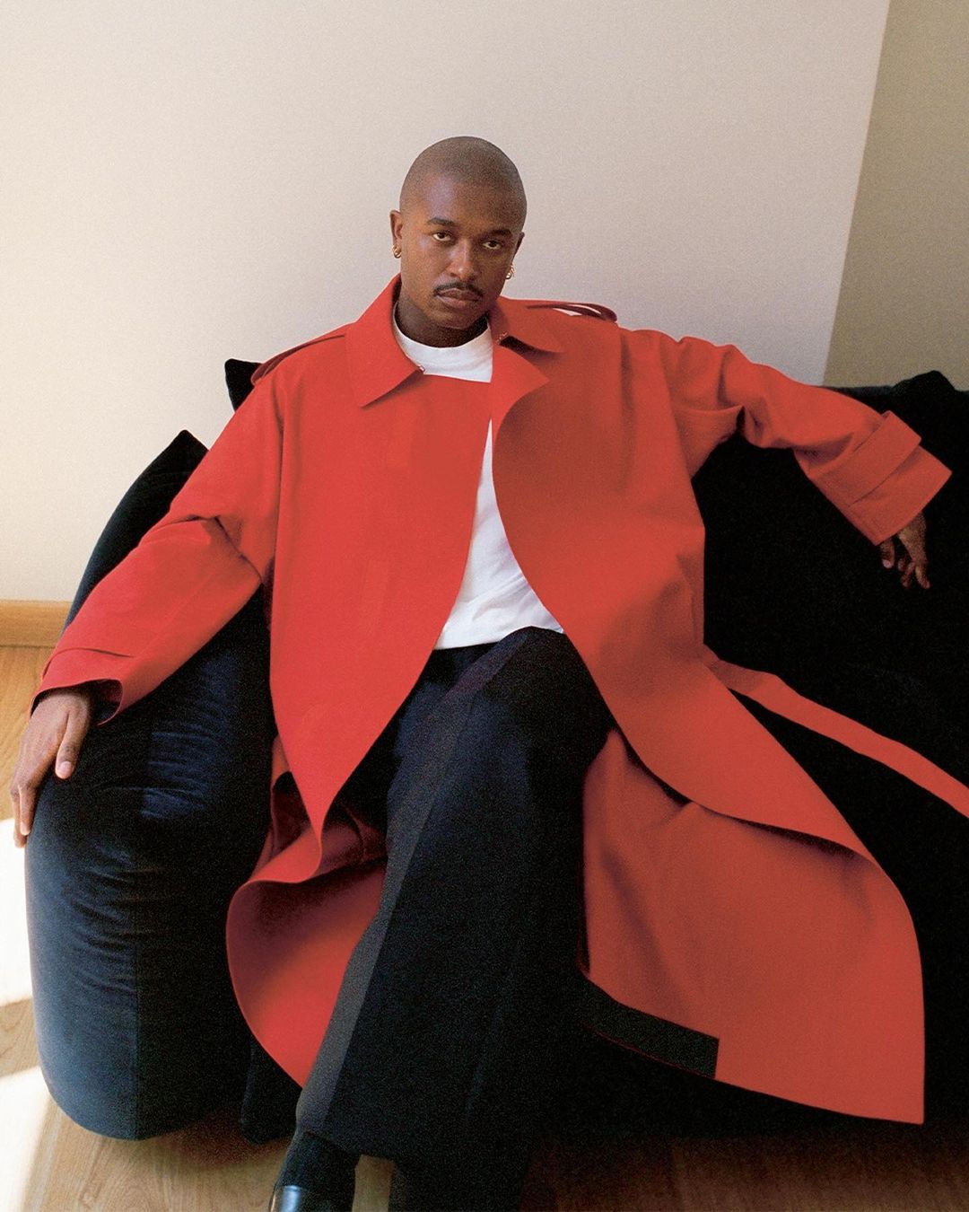 Stylish man in red trench coat poses indoors on a dark sofa, seated against a light wall with natural light, showcasing fashion and portraiture.