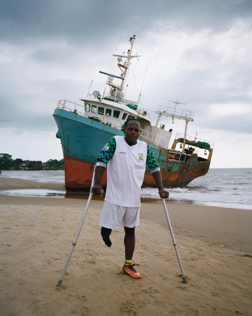 Man with a prosthetic leg standing on a beach with crutches, wearing a soccer uniform, with a large abandoned ship in the background.