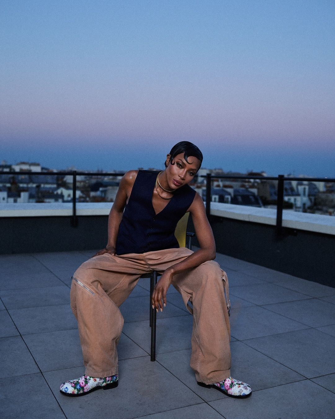 Stylish adult posing on a rooftop wearing vest, trousers, floral-pattern shoes with a cityscape and dusk sky in the background.