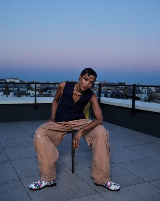 Stylish adult posing on a rooftop wearing vest, trousers, floral-pattern shoes with a cityscape and dusk sky in the background.