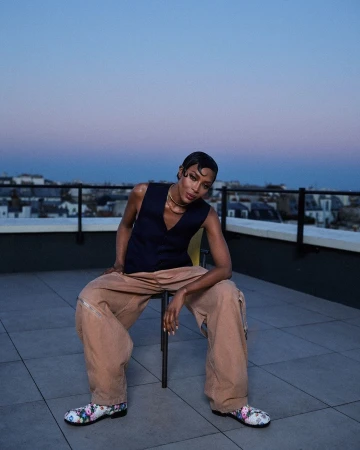 Stylish adult posing on a rooftop wearing vest, trousers, floral-pattern shoes with a cityscape and dusk sky in the background.