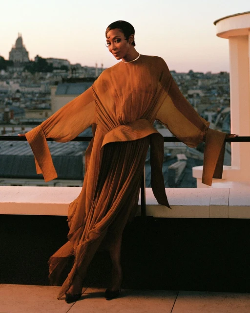 Elegant woman poses in flowing dress on a rooftop with a city view at dusk. The model is wearing a dress, and the scene is serene.