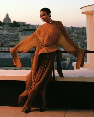 Elegant woman poses in flowing dress on a rooftop with a city view at dusk. The model is wearing a dress, and the scene is serene.