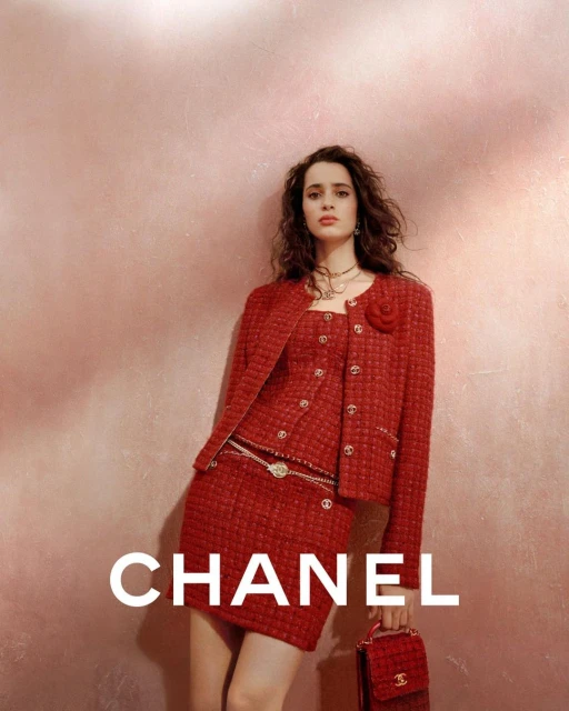 Elegant woman in a red tweed Chanel skirt suit with a matching purse, posing against a pink textured background for a fashion editorial.