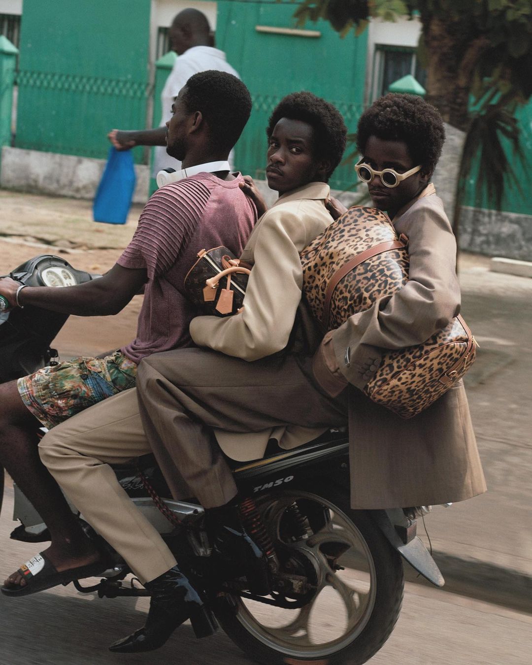 Three Black men on a scooter in an urban environment. They are carrying handbags and wearing fashionable clothing. A sunny, outdoor scene.