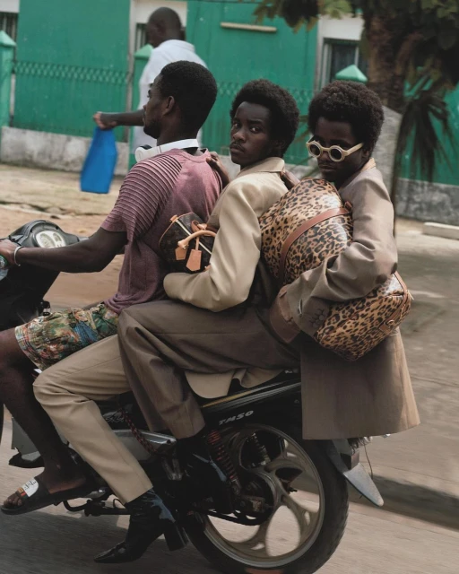 Three Black men on a scooter in an urban environment. They are carrying handbags and wearing fashionable clothing. A sunny, outdoor scene.