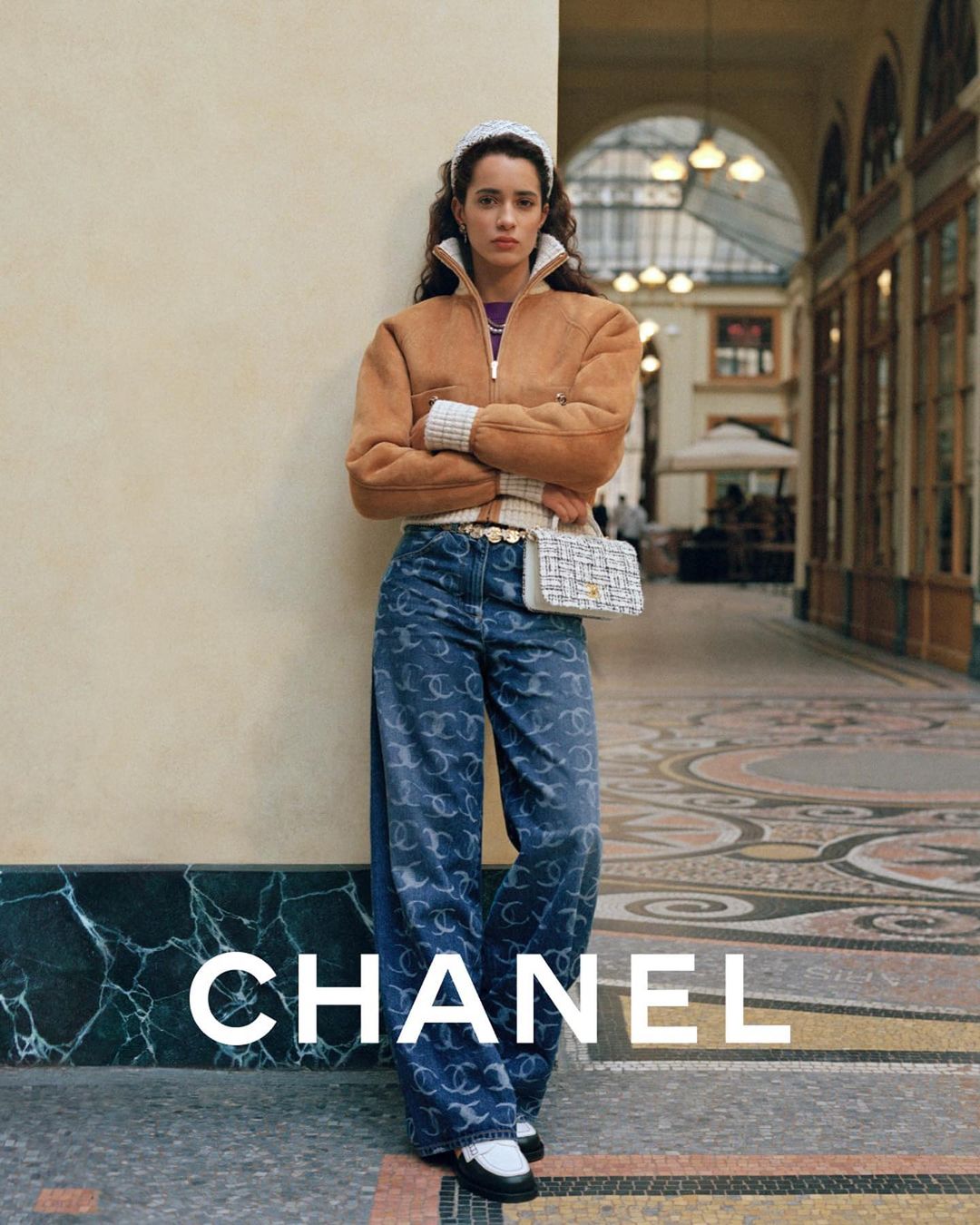 Woman in Chanel outfit posing in an arcade, featuring a tan bomber jacket, logo-patterned jeans, a woven handbag, and black and white loafers.