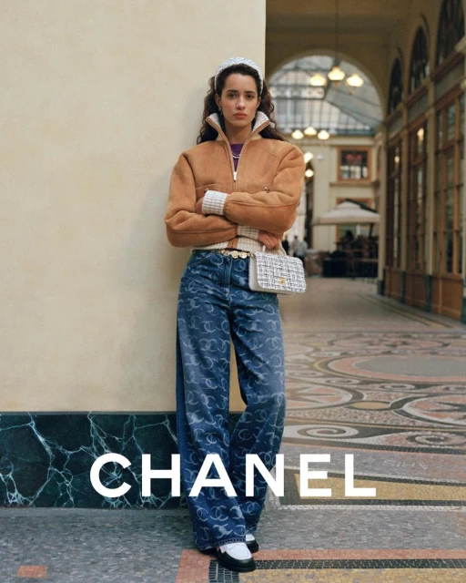 Woman in Chanel outfit posing in an arcade, featuring a tan bomber jacket, logo-patterned jeans, a woven handbag, and black and white loafers.