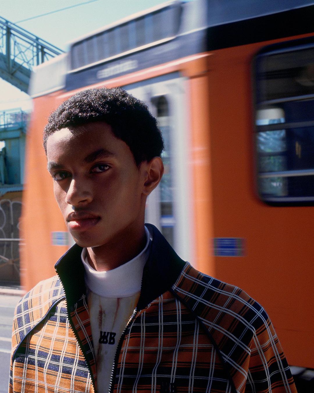 Portrait of a young Black man in a plaid jacket, with a blurred train moving in the background in an urban setting.