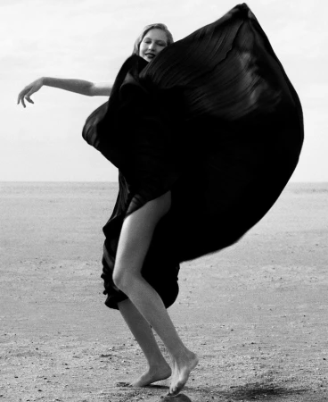 Black and white portrait of a woman in a flowing dark dress billowing in the wind on a sandy beach.