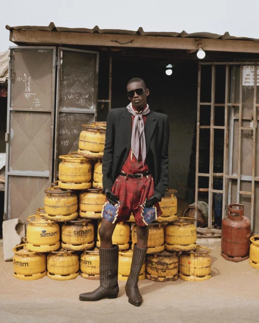A tall, dark-skinned man in shorts, boots, blazer, scarf, and sunglasses poses in front of a shop with stacks of yellow gas tanks.