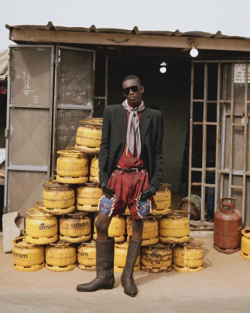 A tall, dark-skinned man in shorts, boots, blazer, scarf, and sunglasses poses in front of a shop with stacks of yellow gas tanks.