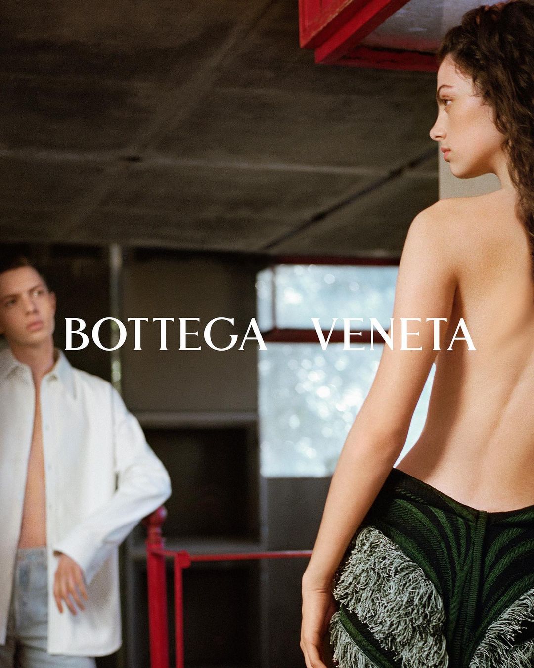 Fashion shot with a woman in green fringe bottoms and a man in a white shirt in a modern, indoor environment with concrete walls.