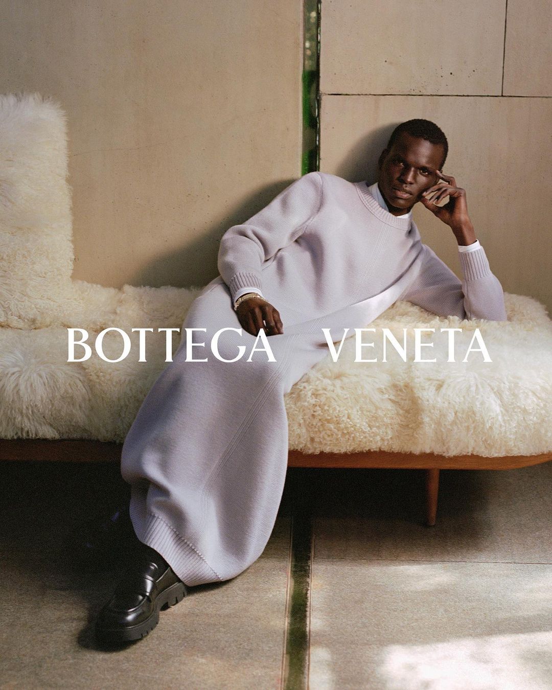 A Black male fashion model poses in a knitwear dress and black shoes while reclining on a fluffy daybed in an indoor setting. 