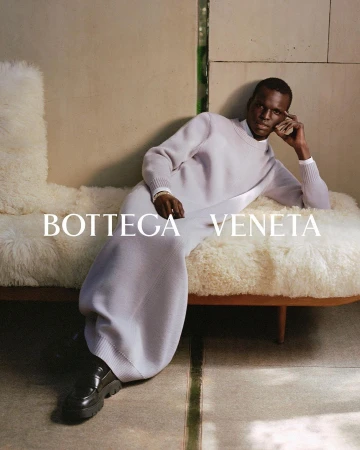 A Black male fashion model poses in a knitwear dress and black shoes while reclining on a fluffy daybed in an indoor setting. 