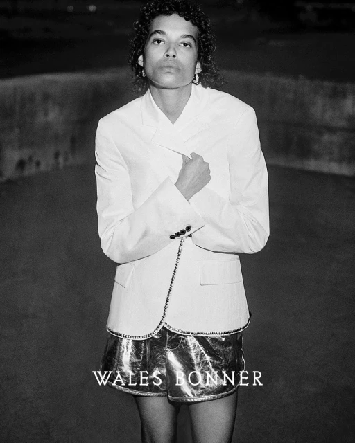 Monochrome portrait of an adult with curly hair wearing a white blazer and metallic shorts, posing against a dark, blurred background.