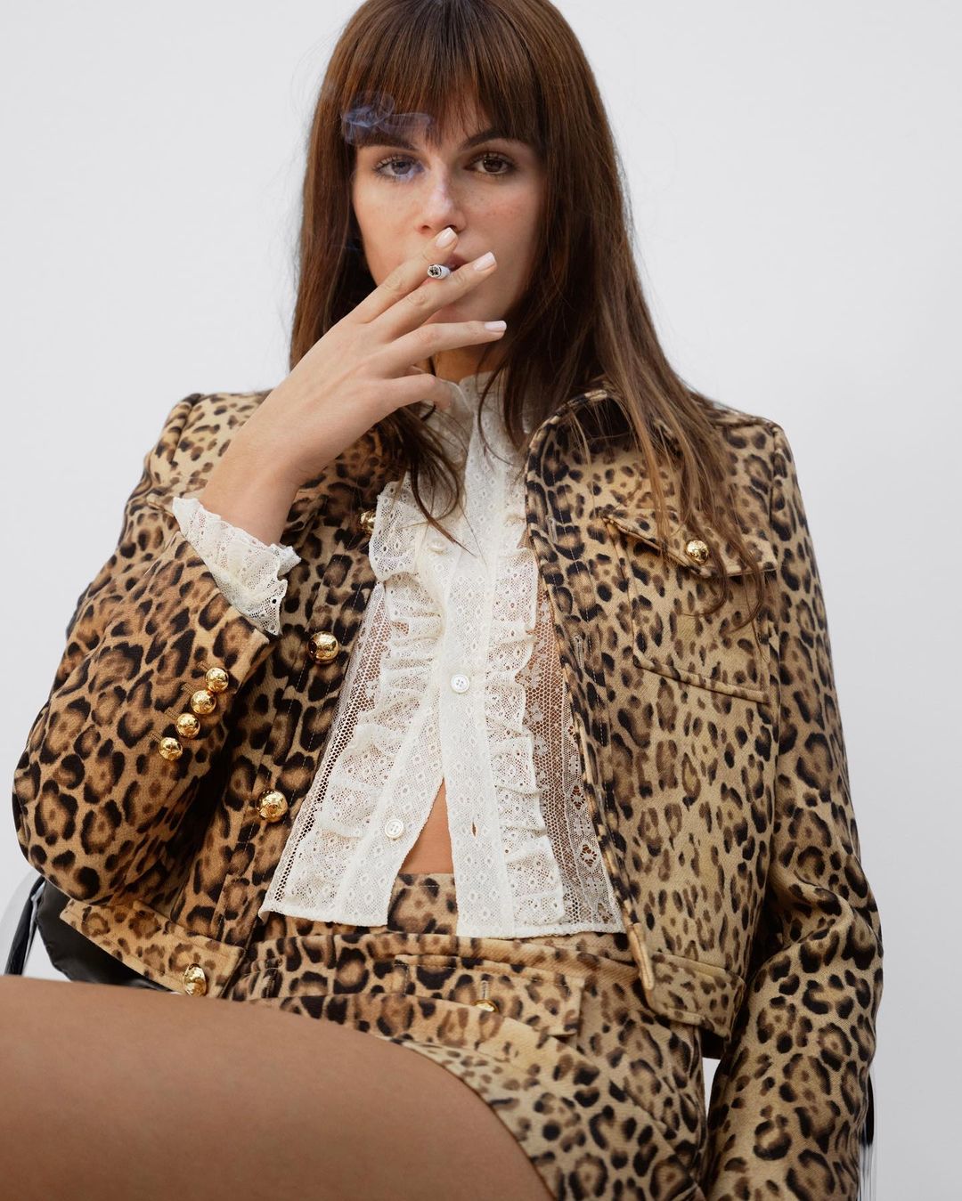 A young adult female with brown hair and bangs sits smoking a cigarette, wearing a leopard print jacket and skirt, in a neutral studio.