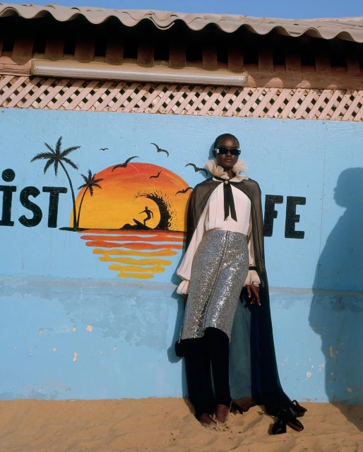 Elegant woman in trendy silver skirt and black cape poses confidently against mural of beach scene with palm trees and surfer.