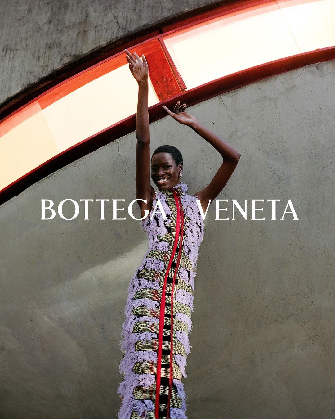 Fashion shot of a Black female model in patterned fringe dress with arms raised against an architectural background. Bottega Veneta logo visible.
