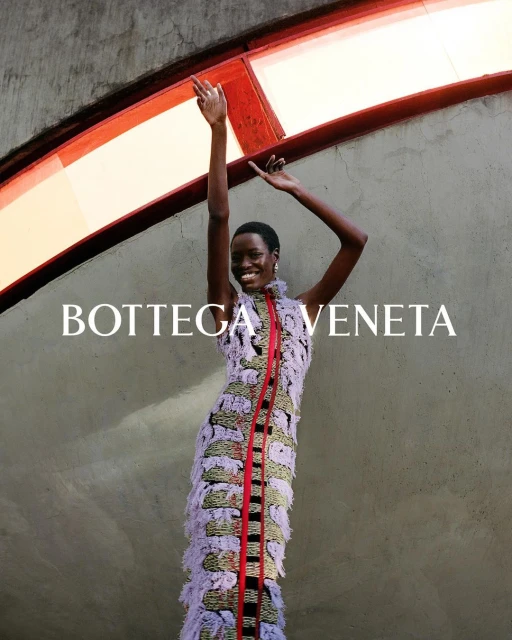 Fashion shot of a Black female model in patterned fringe dress with arms raised against an architectural background. Bottega Veneta logo visible.