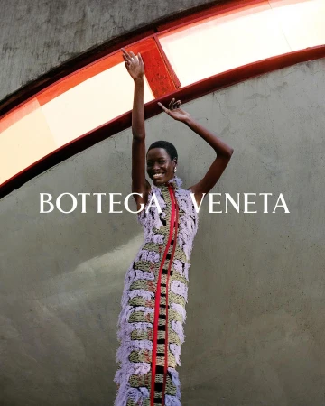 Fashion shot of a Black female model in patterned fringe dress with arms raised against an architectural background. Bottega Veneta logo visible.