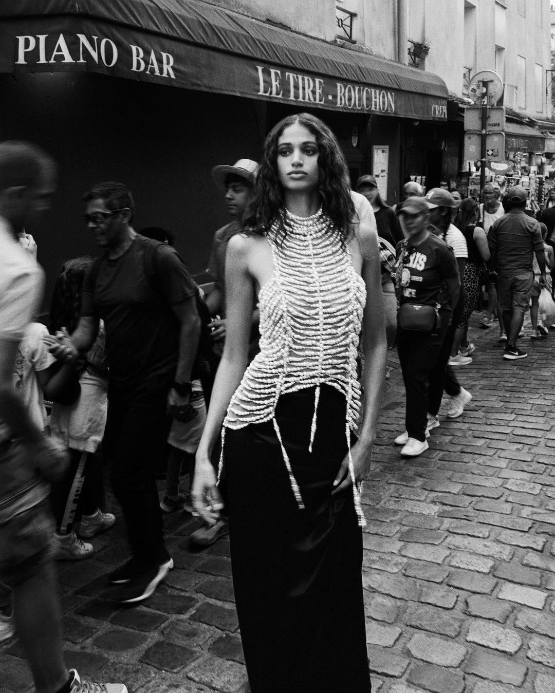 A model poses on a cobblestone street in front of a Parisian bistro, wearing a halter top and long skirt, surrounded by a blurred crowd.