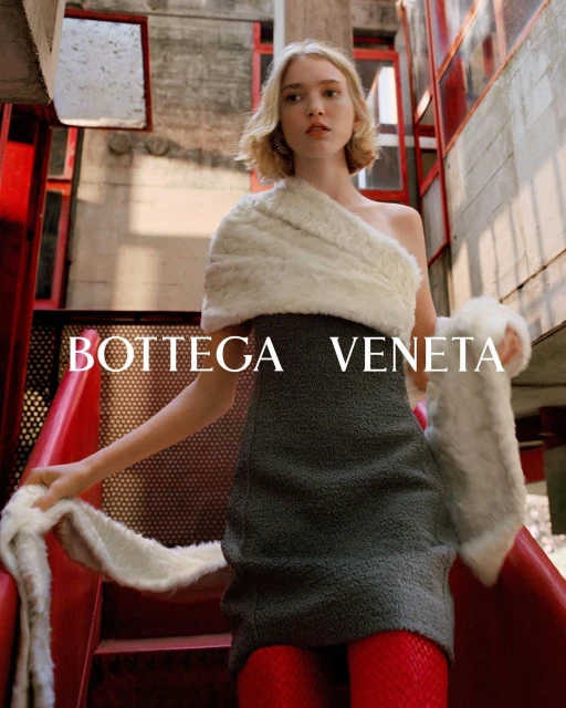 Fashion editorial shot of a young adult woman wearing a grey dress, fur shawl, and red tights, posing on red stairs near a concrete wall.
