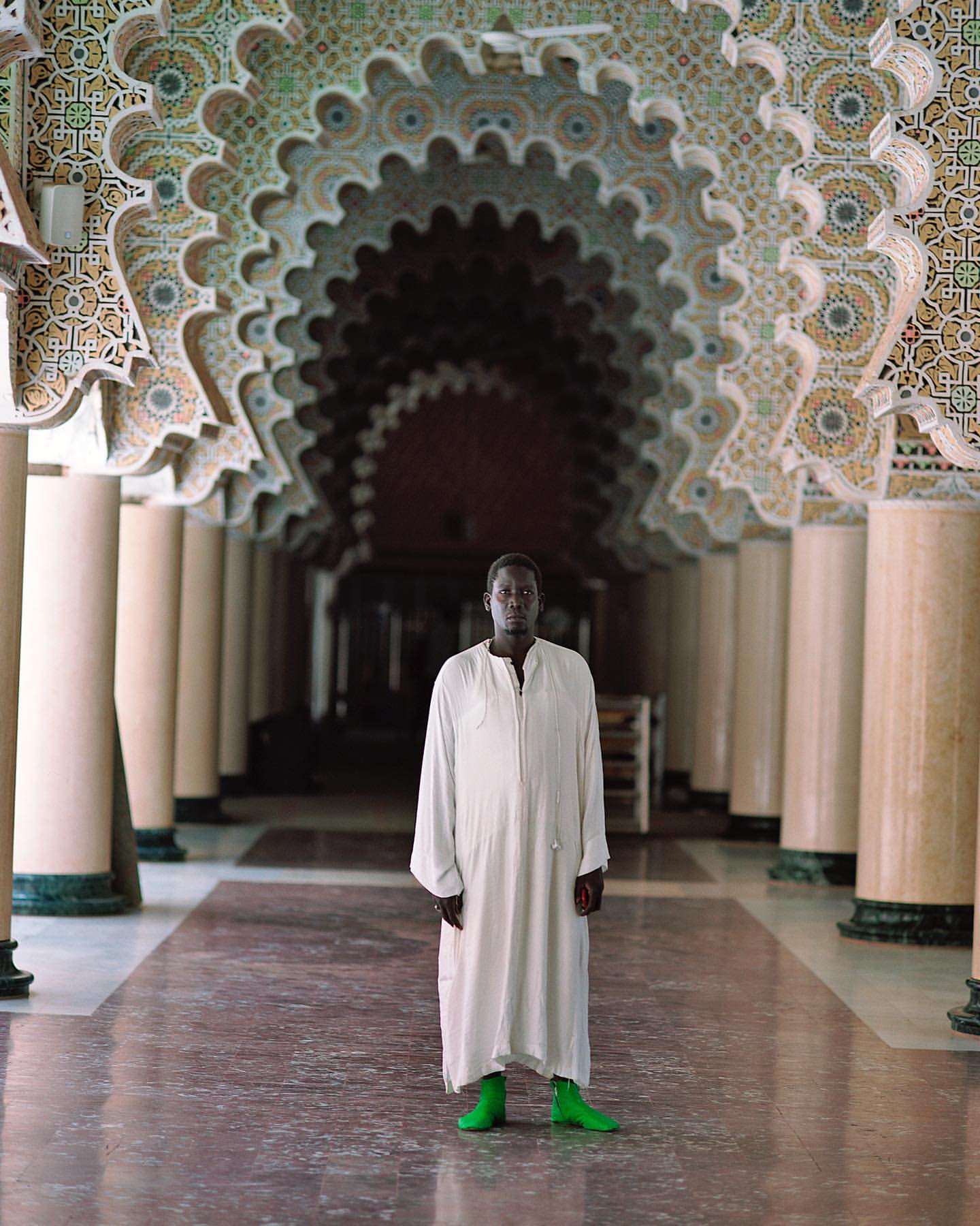 A man stands in a grand hall with decorated arches, wearing a white robe and bright green boots, showcasing architectural details.
