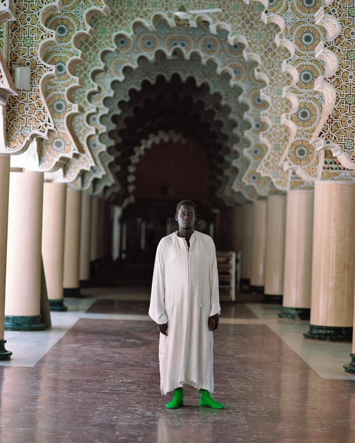 A man stands in a grand hall with decorated arches, wearing a white robe and bright green boots, showcasing architectural details.