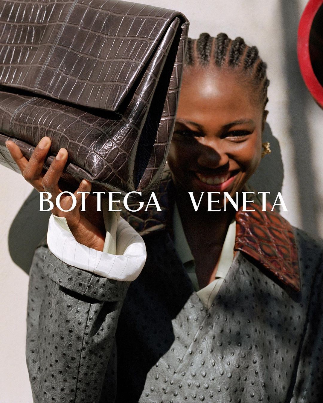 Elegant African woman showcases a brown crocodile embossed handbag and wears a patterned jacket in soft light, with the Bottega Veneta logo.