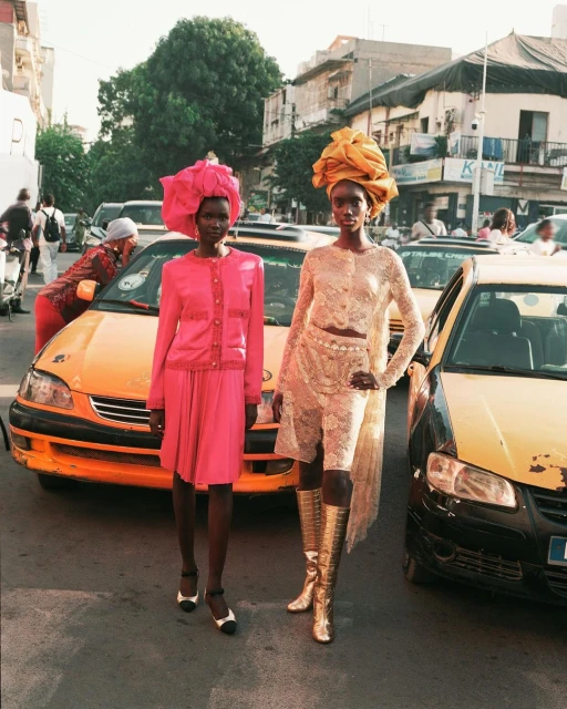 Two African women pose on a city street wearing pink and beige matching outfits and large headwraps, with yellow taxicabs nearby.