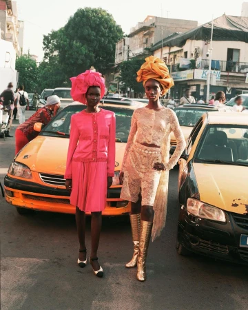 Two African women pose on a city street wearing pink and beige matching outfits and large headwraps, with yellow taxicabs nearby.