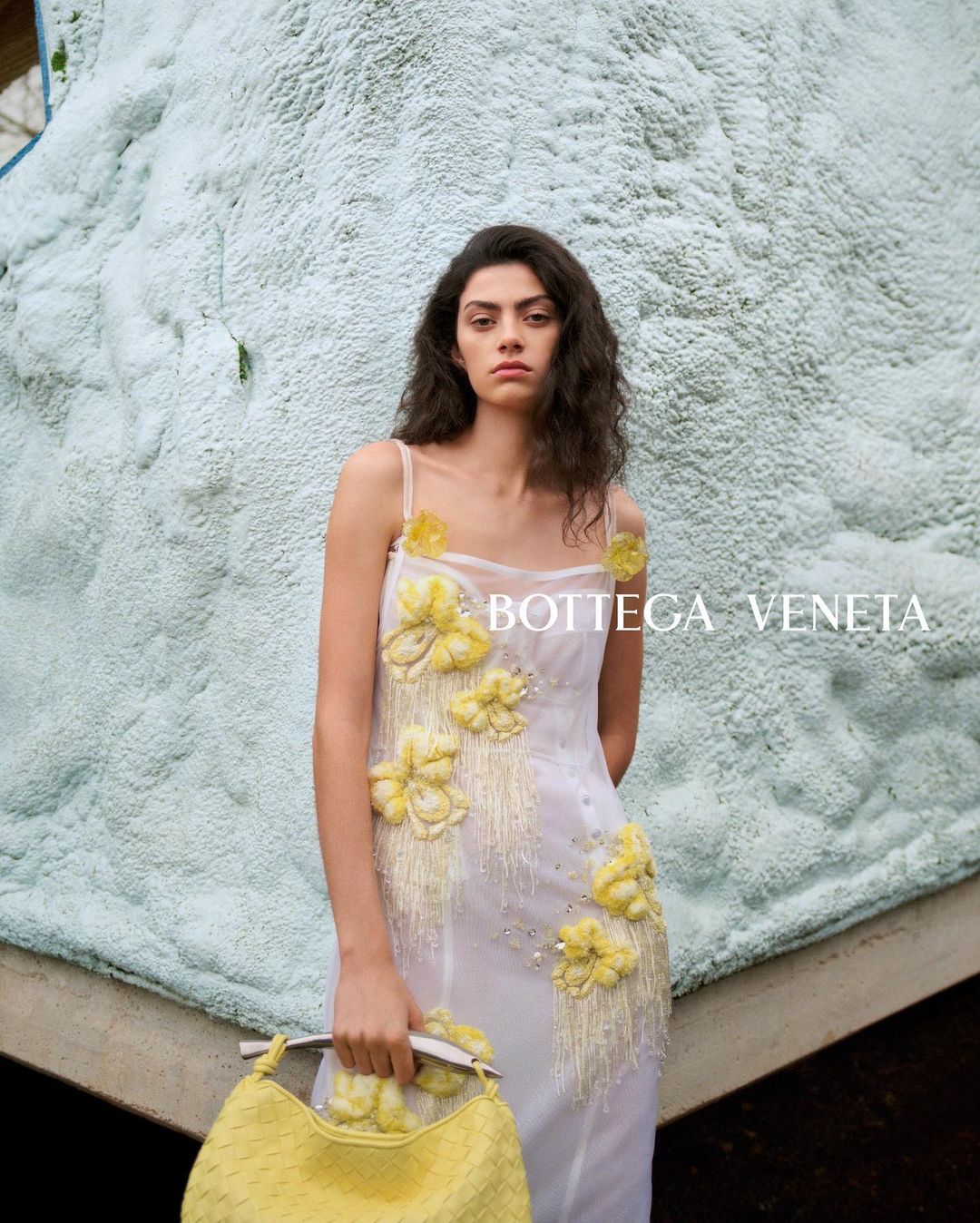 A woman in a white floral dress poses with a yellow woven bag against a light-blue textured wall with natural light. Fashion editorial.