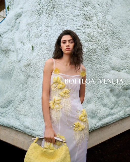 A woman in a white floral dress poses with a yellow woven bag against a light-blue textured wall with natural light. Fashion editorial.