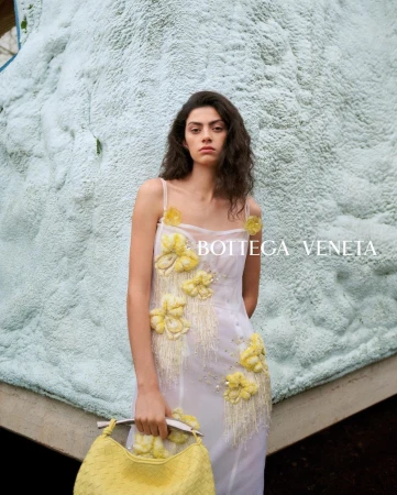 A woman in a white floral dress poses with a yellow woven bag against a light-blue textured wall with natural light. Fashion editorial.