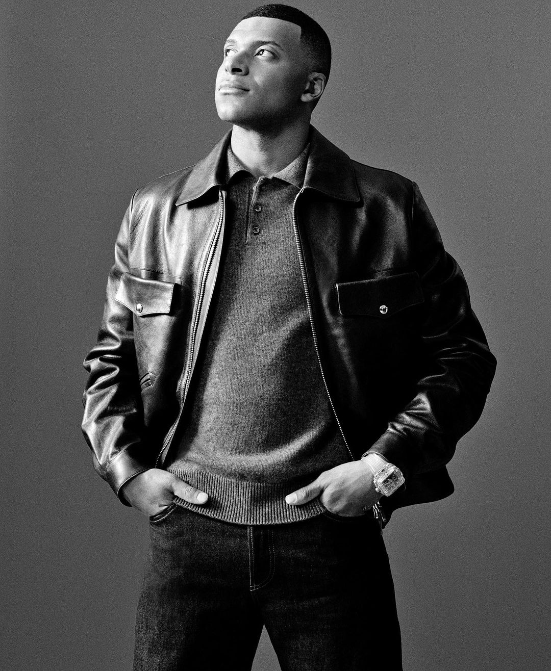 Monochrome portrait of an adult male in a leather jacket and jeans, looking upward with hands in pockets, against a plain gray background.
