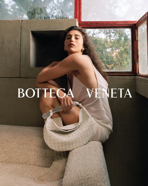 Brunette woman in a white dress poses on concrete steps with a white Bottega Veneta handbag in an indoor setting by a window.