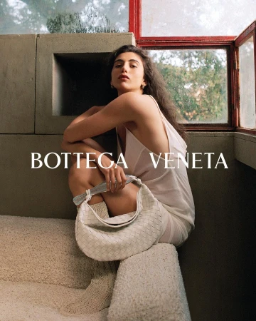 Brunette woman in a white dress poses on concrete steps with a white Bottega Veneta handbag in an indoor setting by a window.