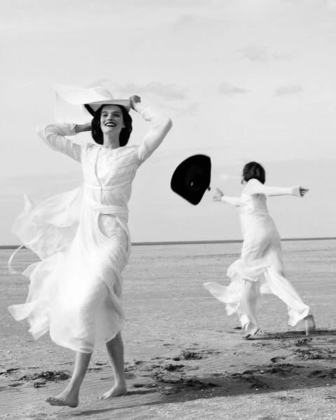 Two women in flowing white dresses play on a sandy beach under a bright sky, with one woman tossing a hat into the air.