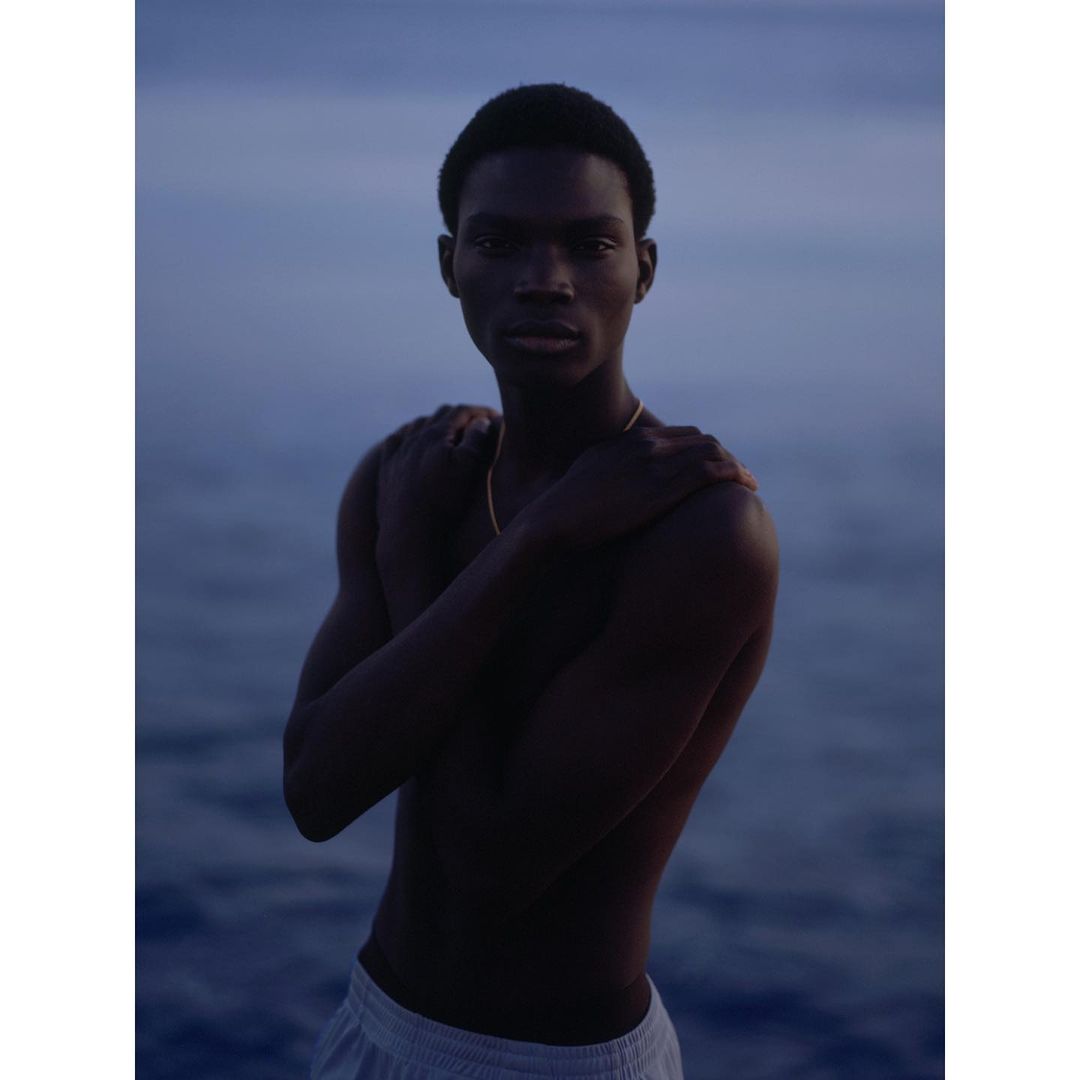 A dark-skinned young adult man with short hair stands with arms crossed in front of a dark, muted blue background, possibly water.