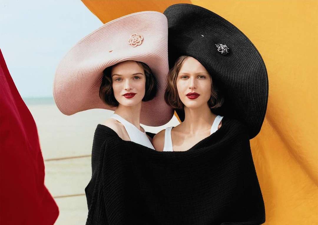 Two women pose in oversized hats and black wraps on a sandy beach with colorful fabric accents, showcasing high fashion.